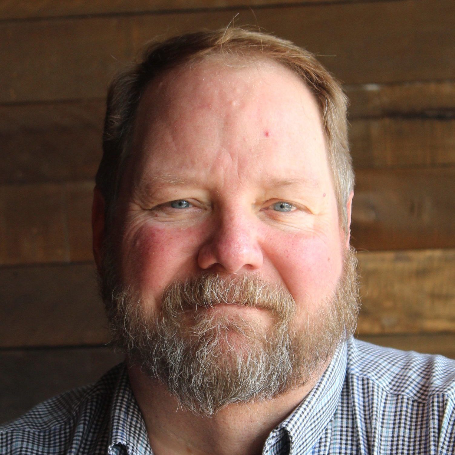 Man with a beard and blue eyes smiling; wearing a blue patterned shirt; set against a wood background.