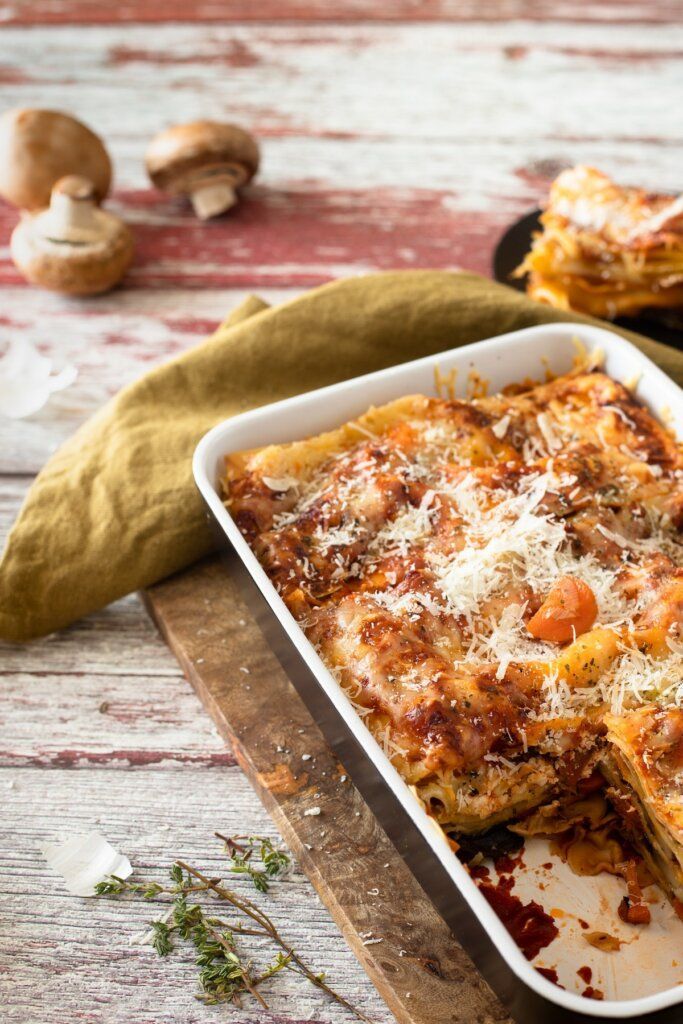 Lasagna in a baking dish on a wooden board, topped with grated cheese; mushrooms and cloth are nearby.