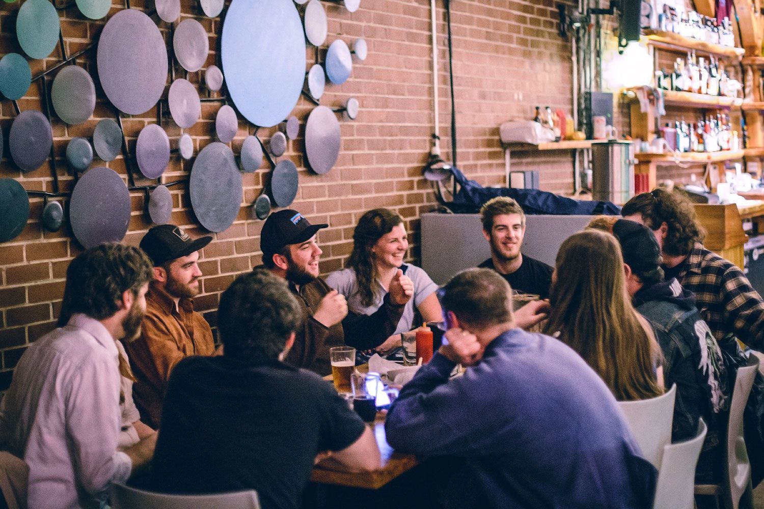 Group of friends at a restaurant table, laughing and talking. Brick wall and decorative circles in background.