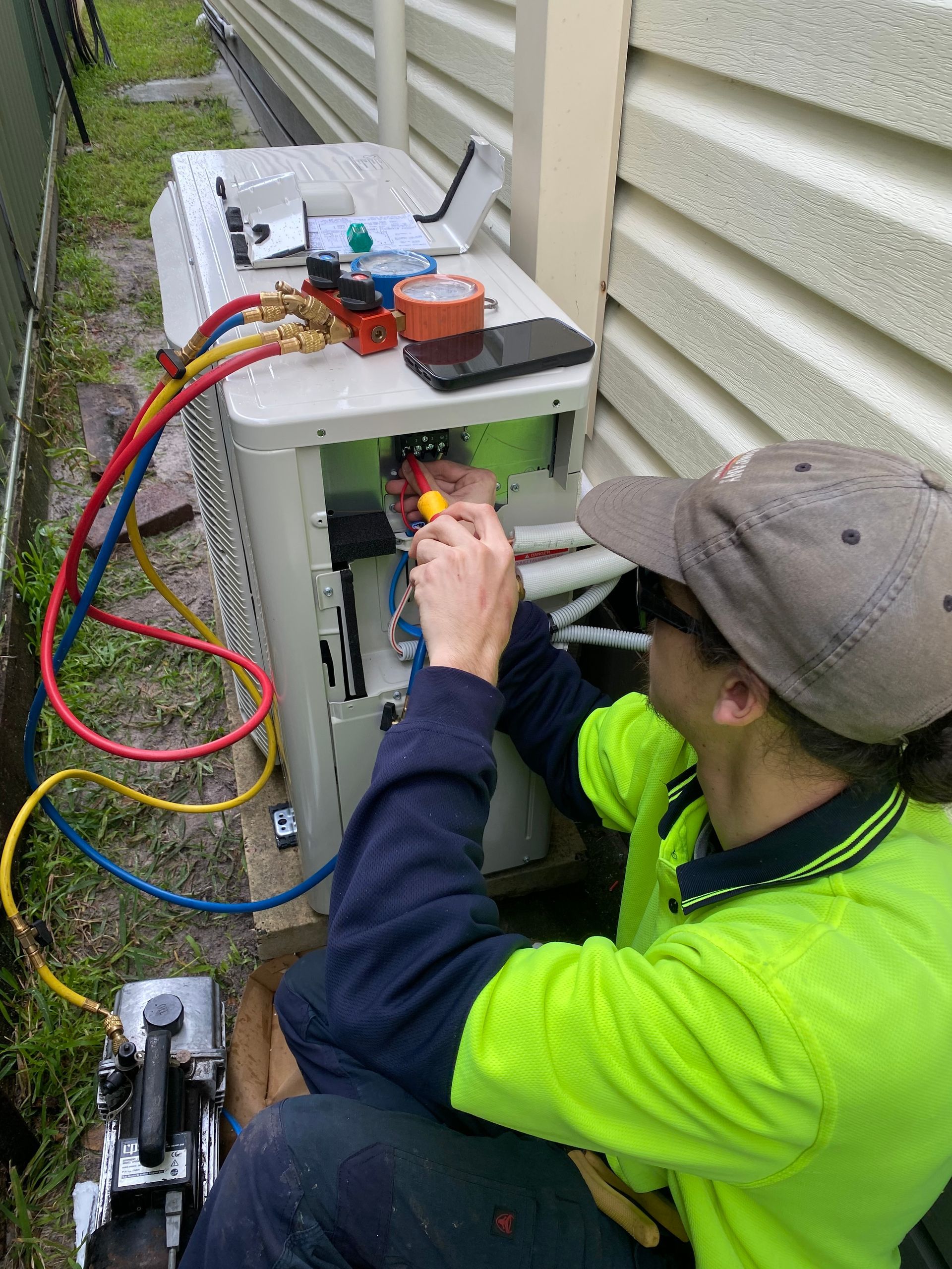 HVAC technician working on an air conditioning unit outside, with gauges, tools, and colored hoses connected. — Airweld Services Pty Ltd in Ulladulla, NSW