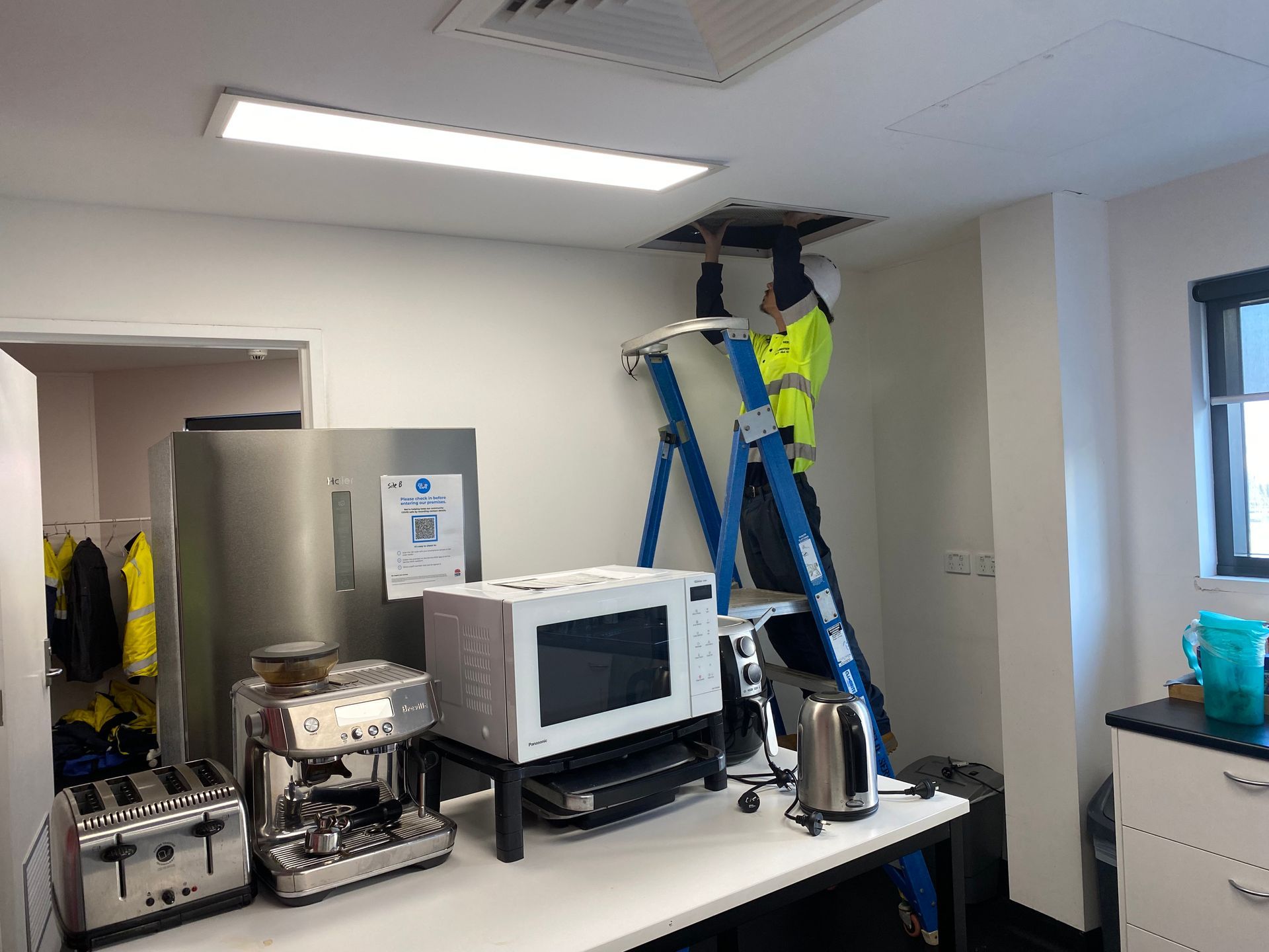 A person in high-vis vest on a ladder, working on a ceiling panel in an office kitchenette. — Airweld Services Pty Ltd in Nowra, NSW