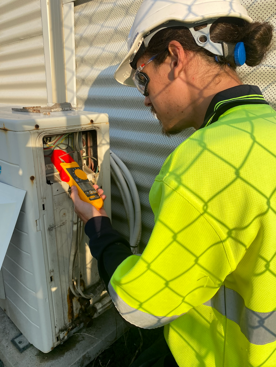 Person in a neon safety vest checks an air conditioner with a multimeter outside — Airweld Services Pty Ltd in Shellharbour, NSW