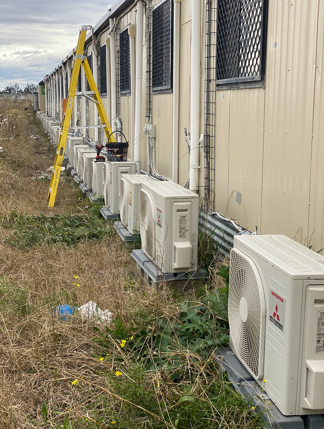 A row of outdoor air conditioning units outside a beige building with a ladder leaning against it — Airweld Services Pty Ltd in Shellharbour, NSW