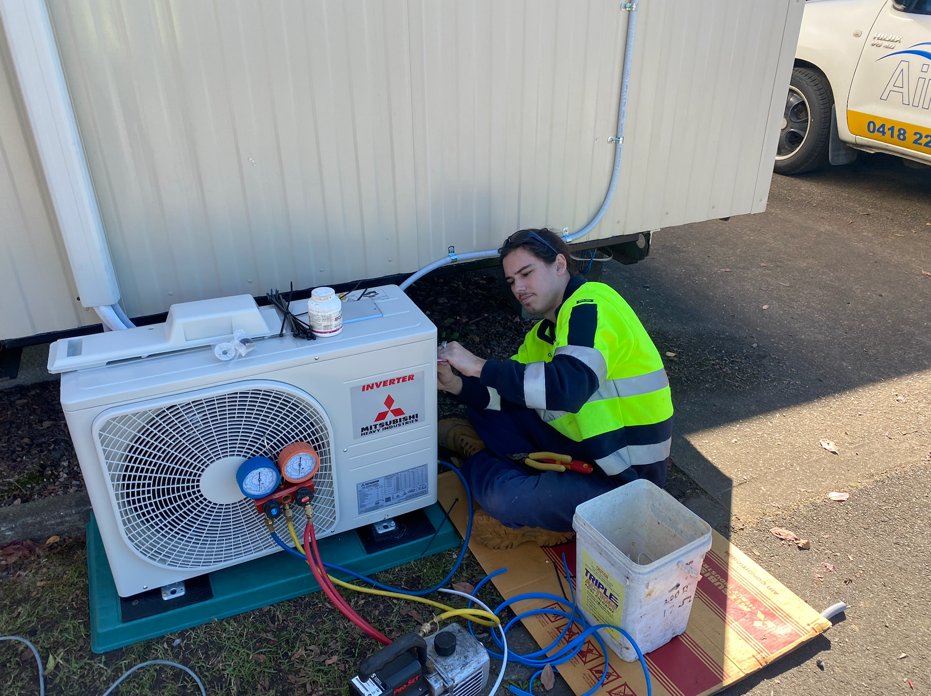Technician repairs an outdoor Mitsubishi AC unit, kneeling, wearing a safety vest, next to a trailer  — Airweld Services Pty Ltd in Wollongong, NSW