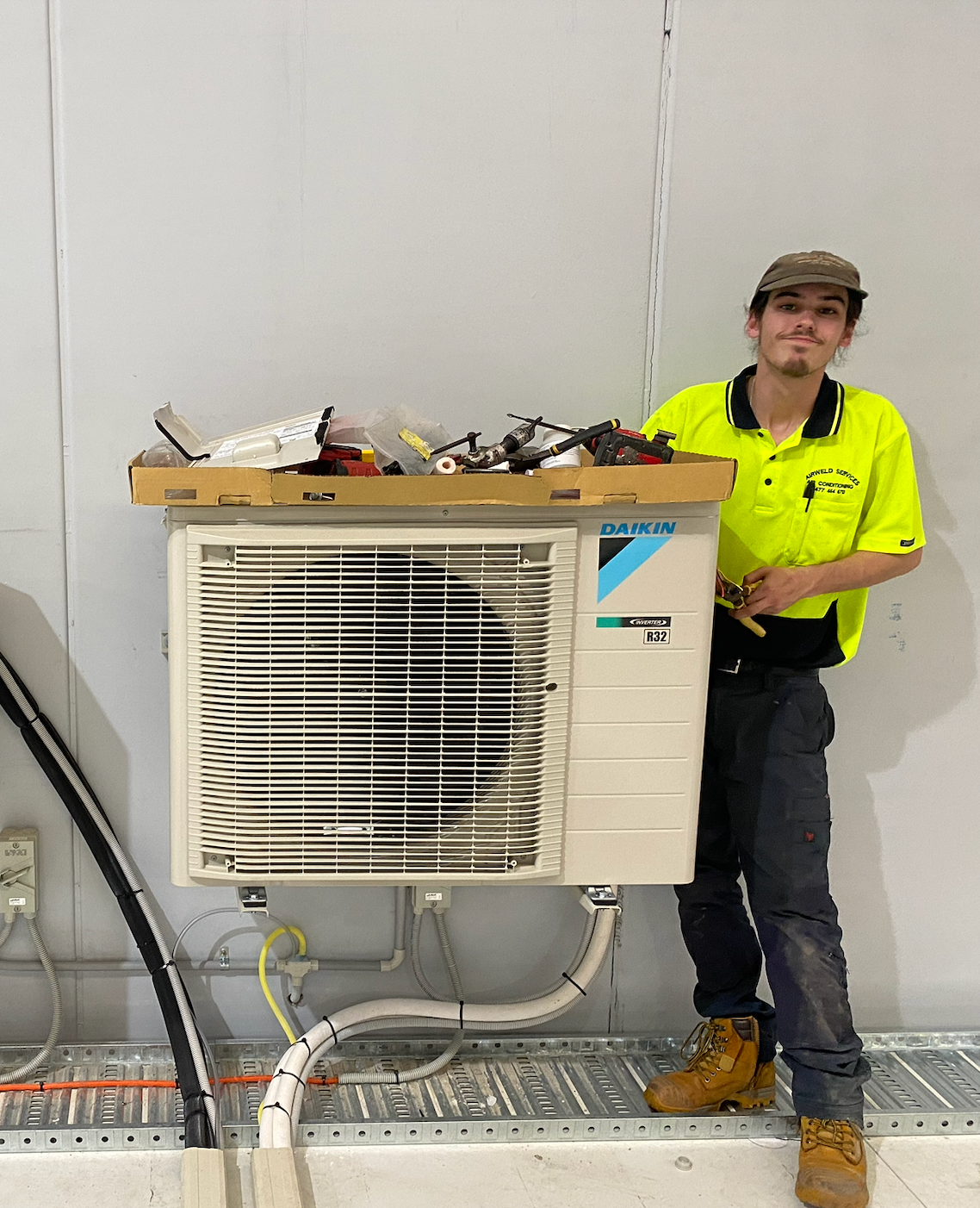 Person standing by an air conditioning unit; holding tools; wearing work clothes; indoors.  — Airweld Services Pty Ltd in Wollongong, NSW