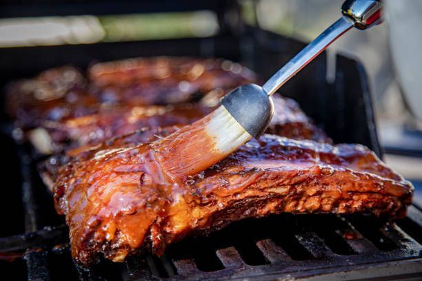 A person is applying sauce to ribs on a grill.