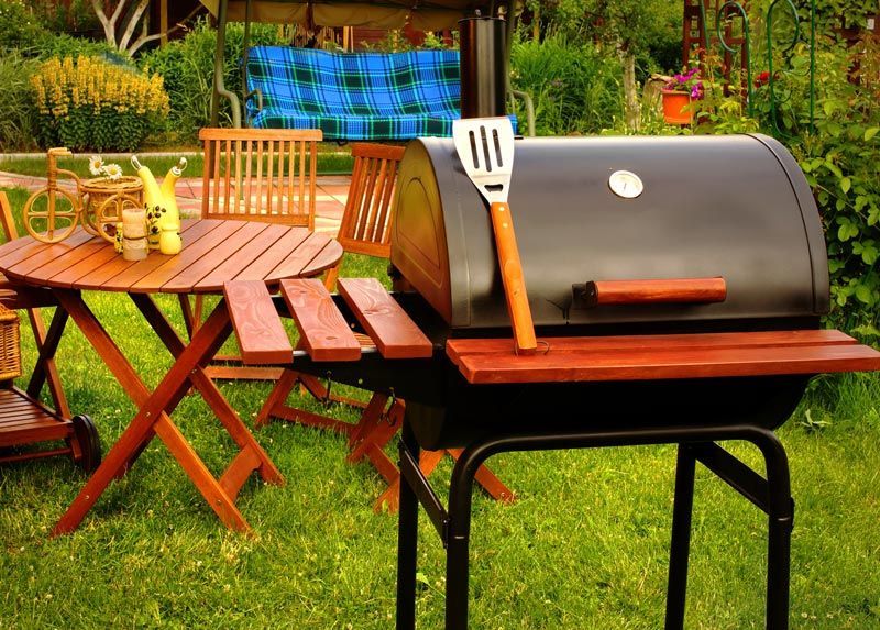 A barbecue grill is sitting in the grass next to a wooden table and chairs.