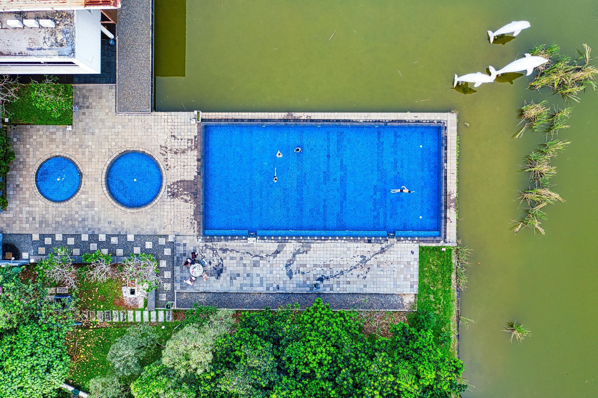 Aerial view of a rectangular blue swimming pool, two smaller round pools, and a lake. People swim in the main pool.