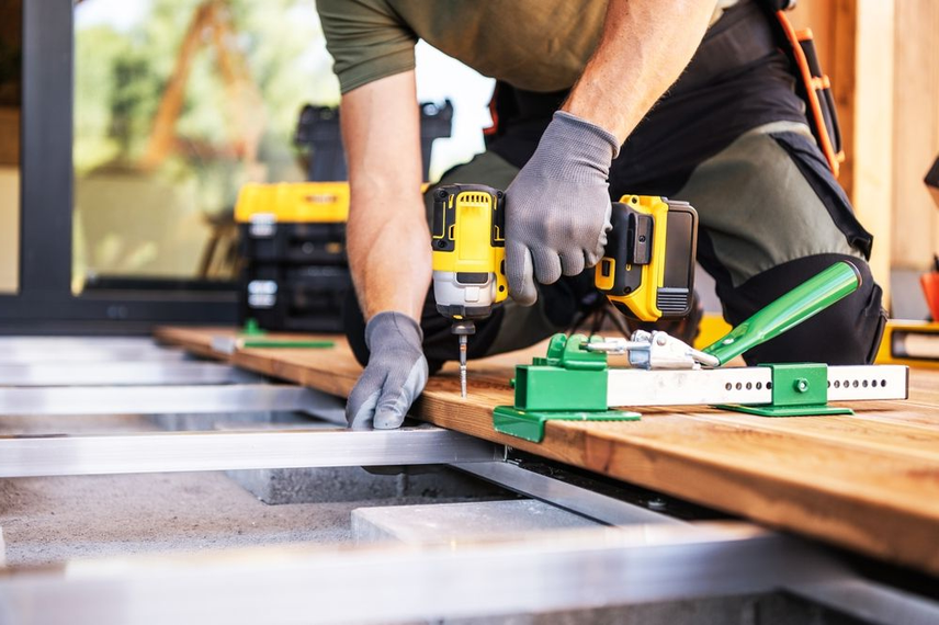Person using a power drill to install wooden deck boards. Gray gloves, tools, and green spacers.