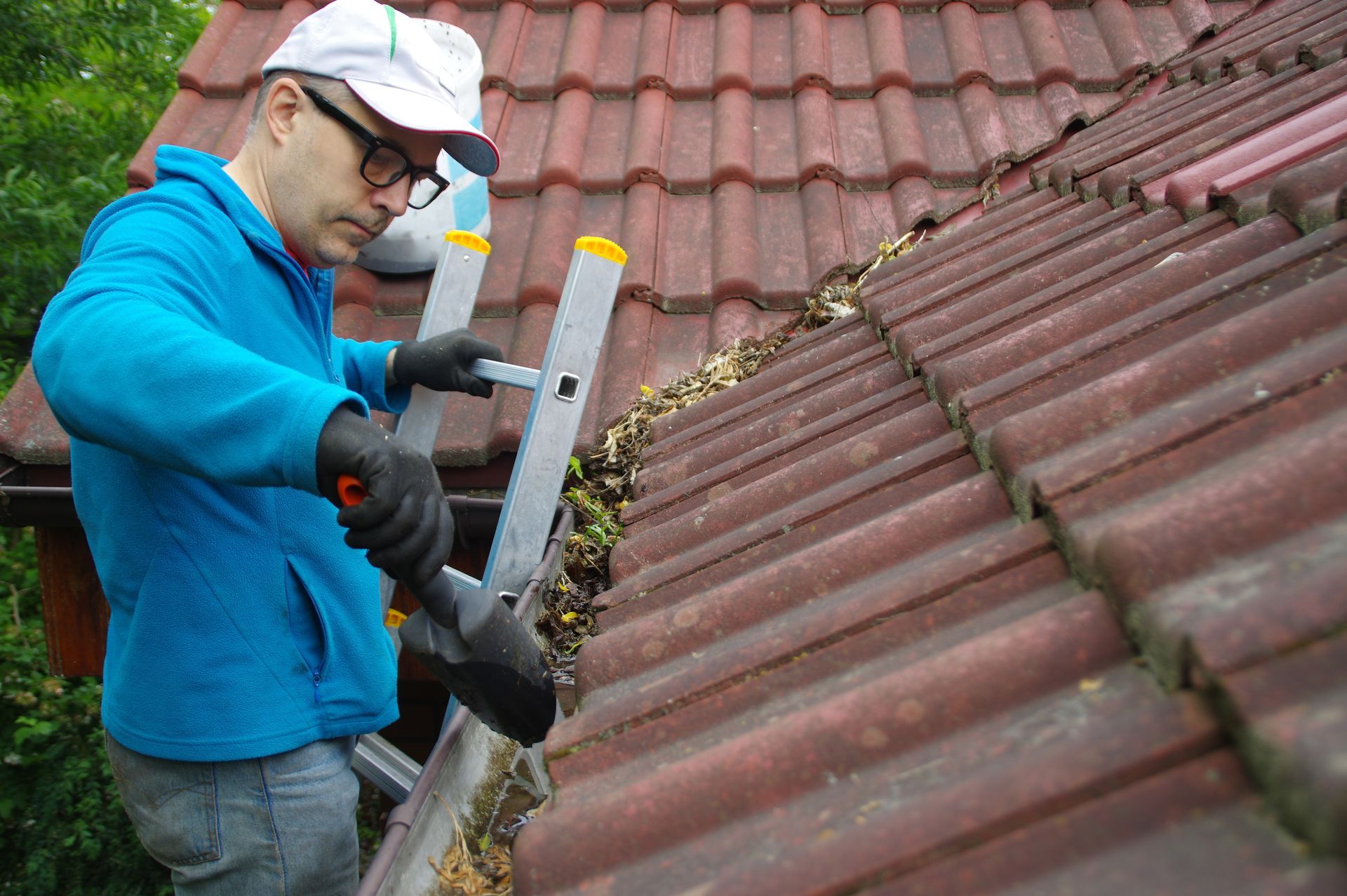 Man on a ladder cleaning leaves from a gutter. He's wearing a blue jacket, gloves, and a white cap, working on a red tile roof.