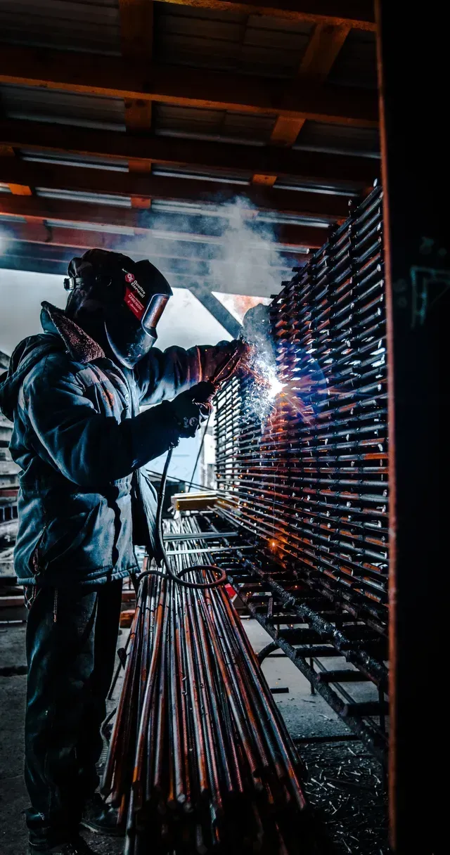 A picture of a man in full welders clothes standing up in a shop and welding rebar at shoulder level with black welders helmet on. He is using a TIG welder. There is a stack of rebar on a rack in front of him and he is slightly leaning over it.