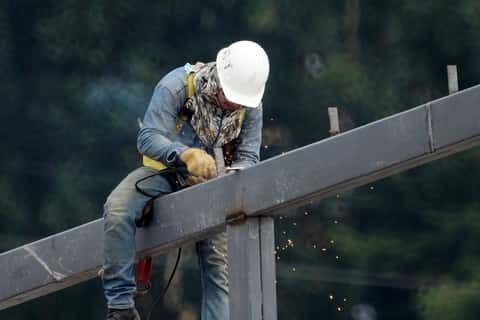 A man up on a steel structural frame with his head down, straddling a beam and welding.  Sparks are flying, he is wearing a white hardhat and blue jeans and a blue dirty shirt.