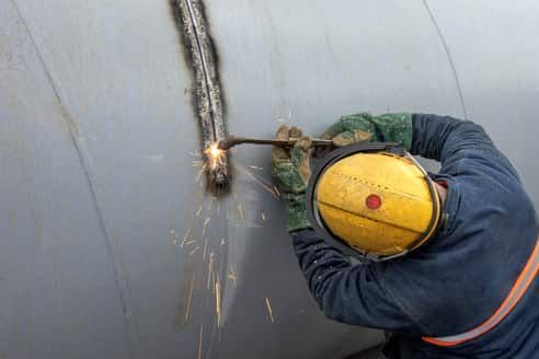 An overhead picture of a man welding a long bead on a giant light gray pipe.  He has on a yellow hard hat, blue jump suit and green leather gloves.  He is only visible from the waist up.