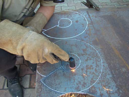 Close up of a person using a hand piece for a plasma cutter to cut sheet steel. The steel sheet has spiral patterns drawn on it with white chalk. The person is cutting following the lines.