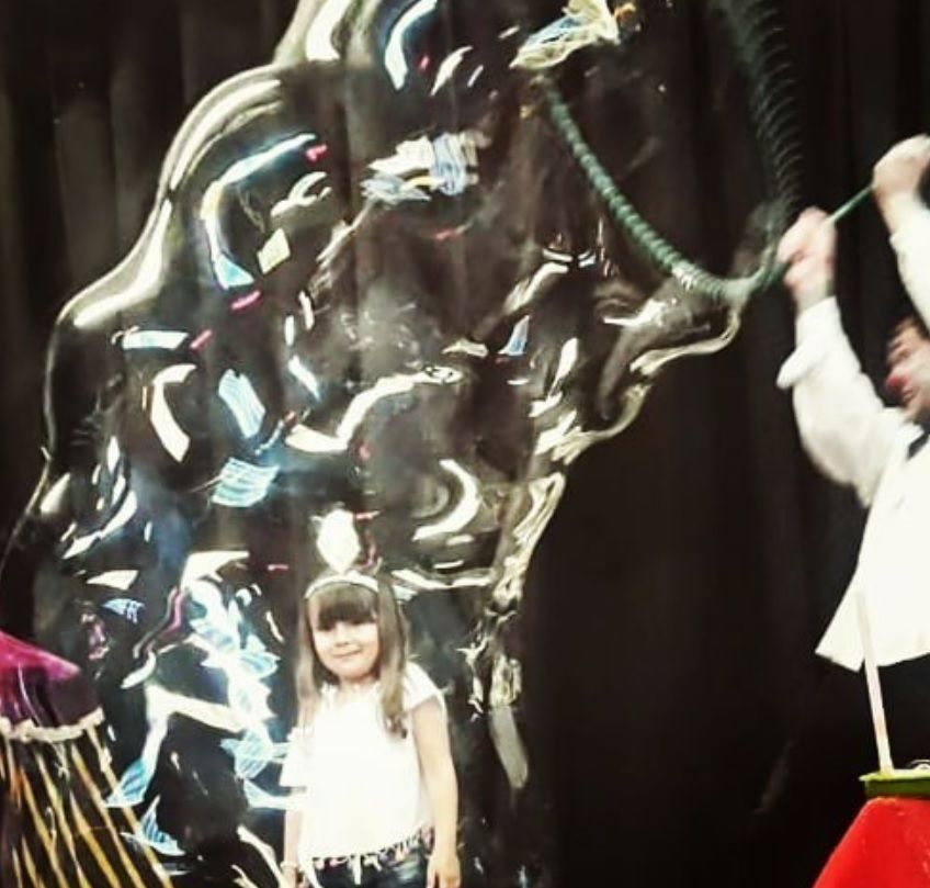 Girl inside giant bubble; performer using wand. Stage background, dark.