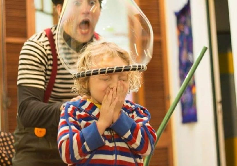 A child inside a large bubble, eyes closed, hands clasped. A person behind them blows the bubble, mouth open.