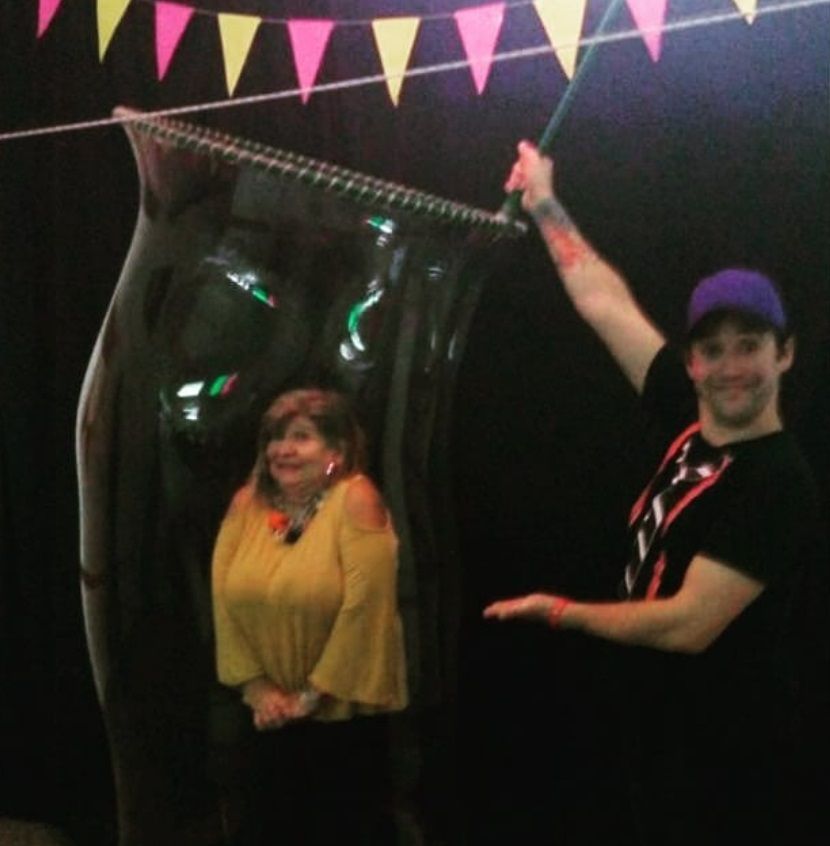 A woman stands inside a large bubble as a man holds the wand to make it. Black background with banners.