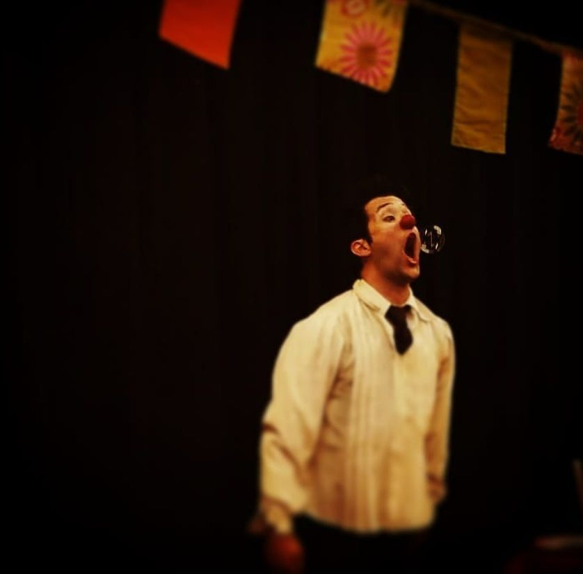 Man blowing a bubble on stage, looking upwards. Black background with colorful banners.