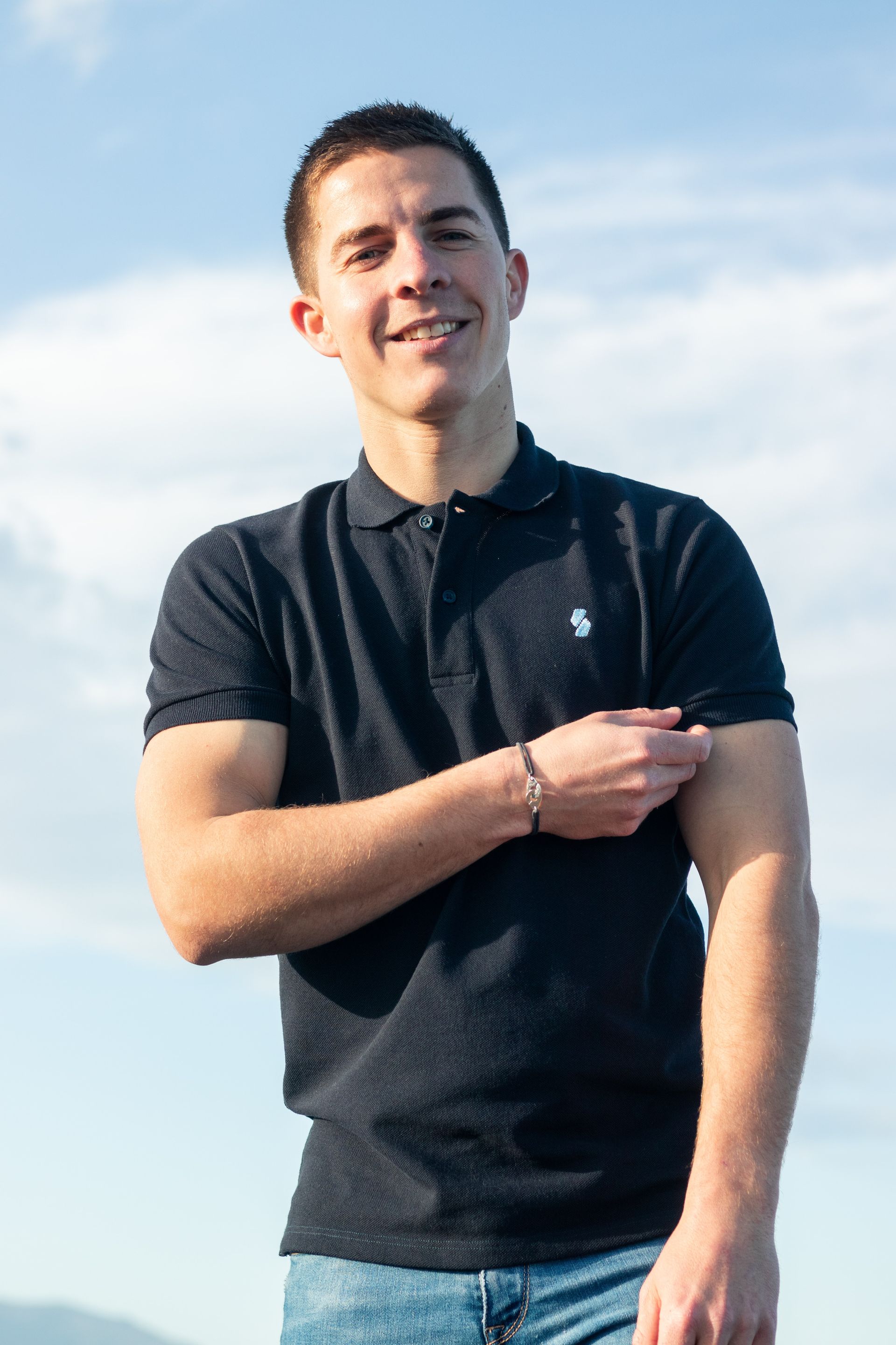 A young man wearing a black polo shirt is standing with his arms crossed and smiling.