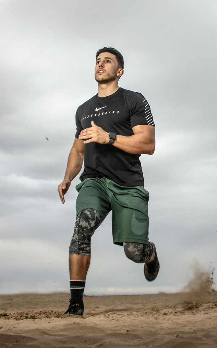 Man running outdoors, wearing black shirt and green shorts, dust cloud around foot, overcast sky.
