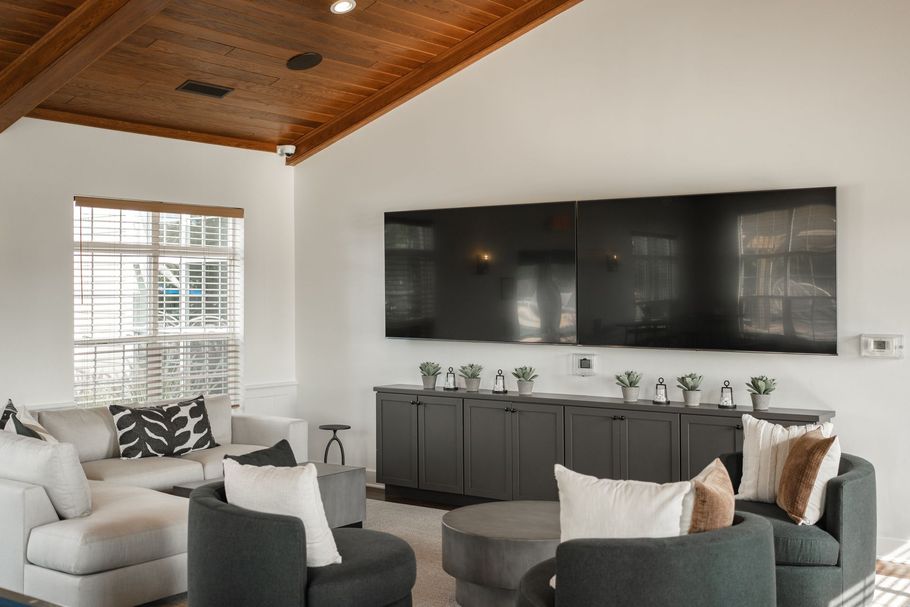 Living room with gray furniture, large TVs, wooden ceiling, and window with blinds.