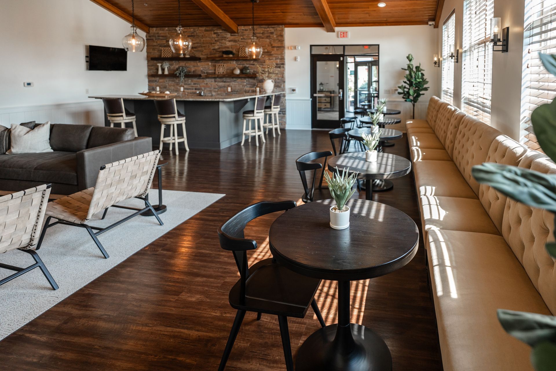 Lounge area with seating, bar, tables, and a brick wall. Bright with sunlight, wooden floors, and modern decor.