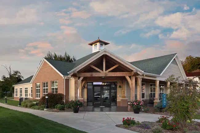 Low-slung brick and wood building with arched entrance, green roof, and small cupola. Paved walkway, green lawn, and flowers in planters.