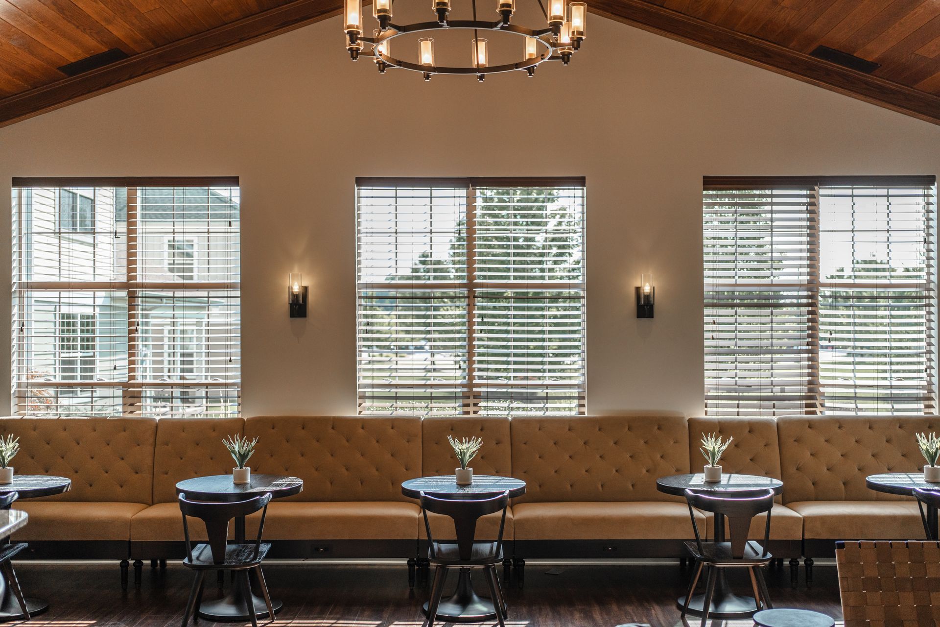 A room with a tan tufted booth along windows, small tables, and a chandelier.