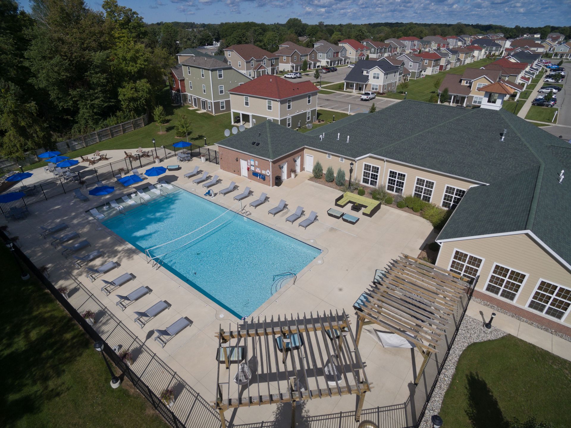 Aerial view of a pool area, beige building with a green roof, and houses in a residential neighborhood.