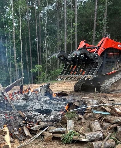 Orange excavator clearing debris beside a smoky burn pile in a forested area