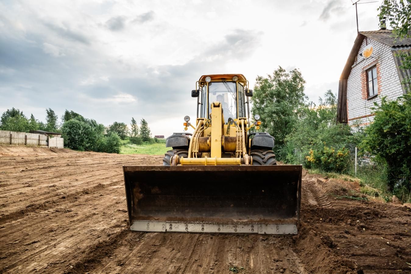 Yellow backhoe grading a dirt lot, with a house and trees in the background under a cloudy sky.
