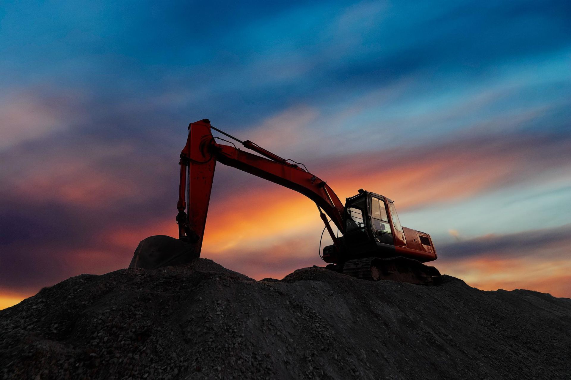An orange excavator silhouetted against a dusk sky, atop a pile of earth.