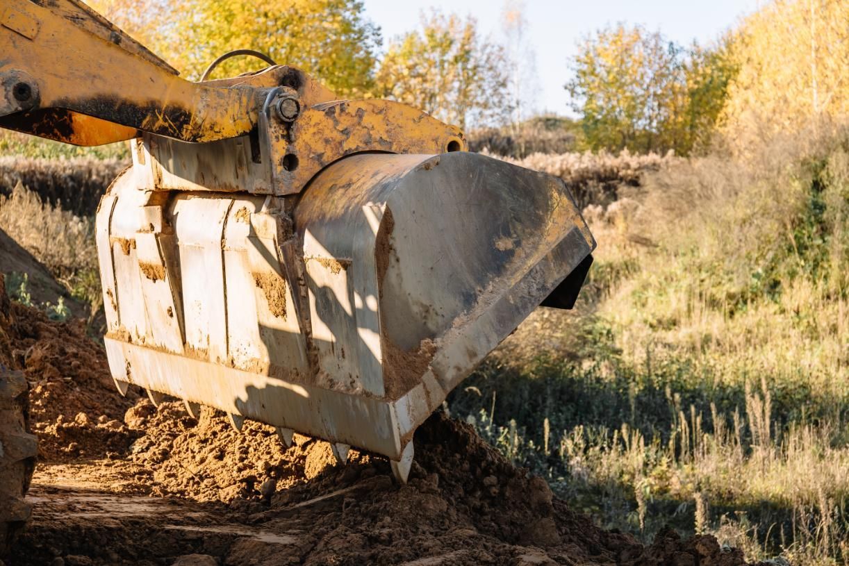 Yellow excavator bucket scooping dirt in a construction site with trees in the background.
