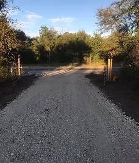 Gravel driveway leading into a wooded area. Wooden posts mark the entrance.
