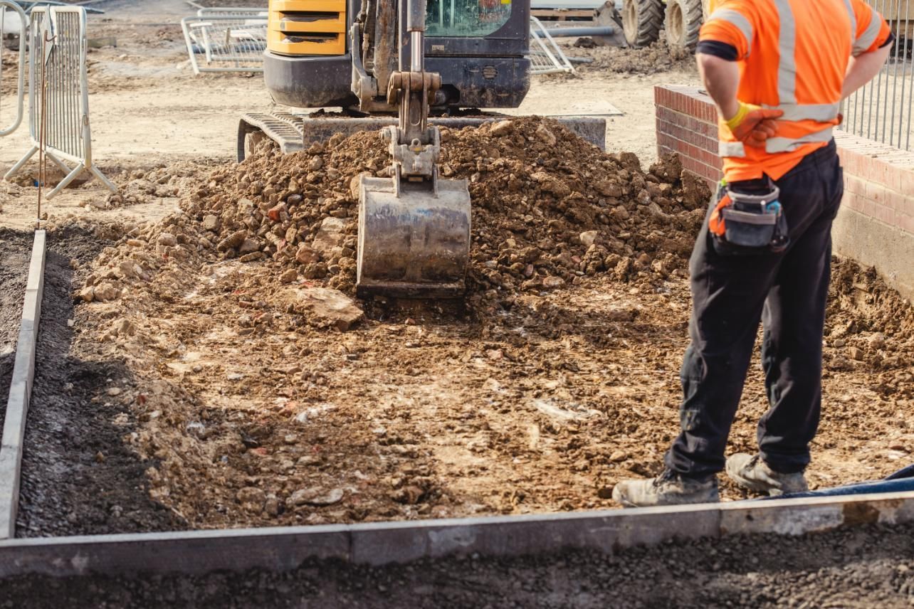 Construction worker in orange vest watches a mini excavator dig dirt at a construction site.