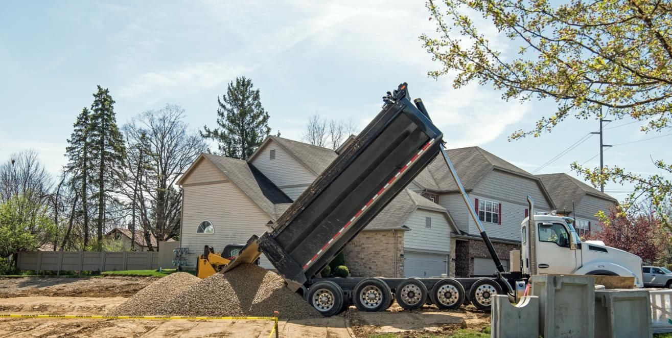 Dump truck unloading gravel in front of a house on a sunny day.