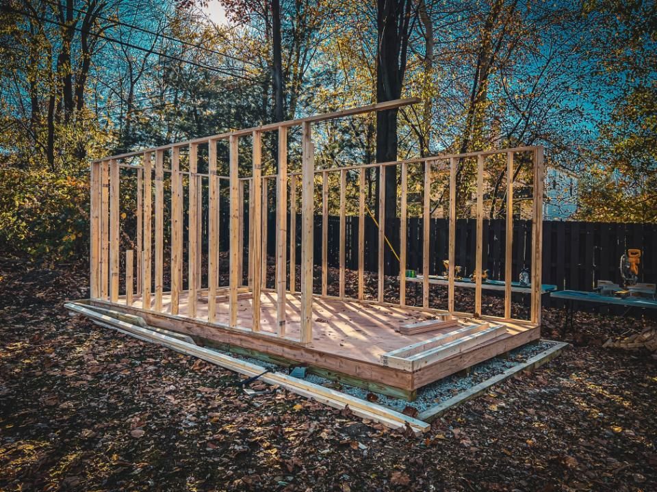Wooden shed frame under construction in a backyard, surrounded by trees and a fence.