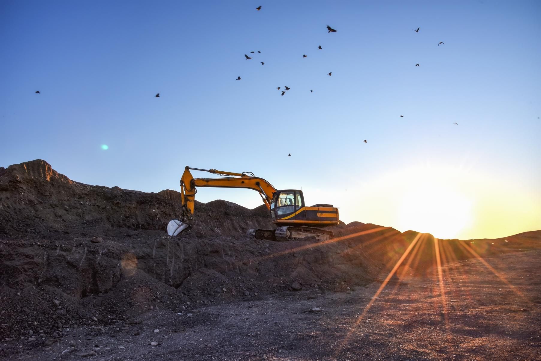 Excavator on a pile of debris at sunset with birds flying overhead.