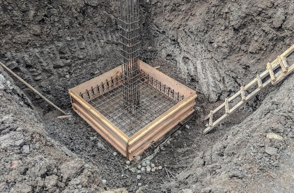 Construction site. Wooden forms encase a square concrete foundation with rebar framework, in a dirt pit.