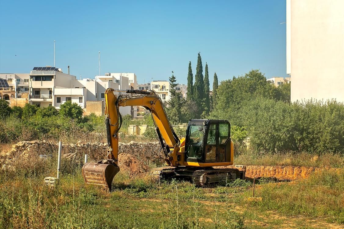 Yellow excavator in a field, clearing land near buildings, under a blue sky.