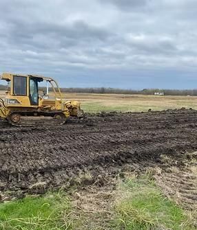 Yellow bulldozer plowing wet, brown field under cloudy sky.