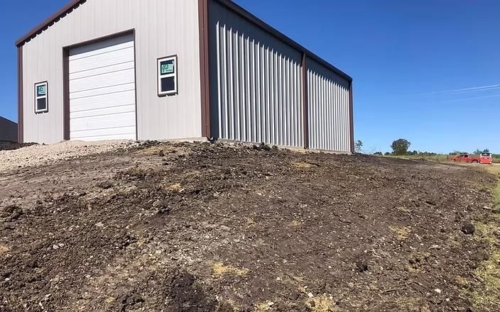 Steel building with a white garage door, brown trim, and a dirt mound in front, under a blue sky.