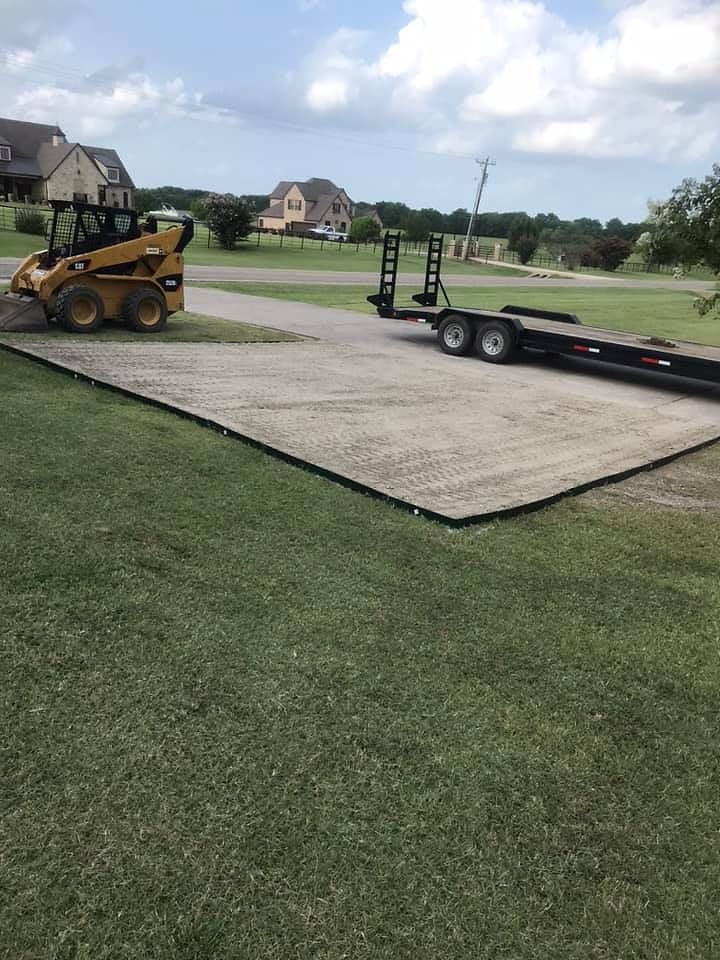 A yellow skid steer and a trailer on a gravel driveway in a grassy yard. 