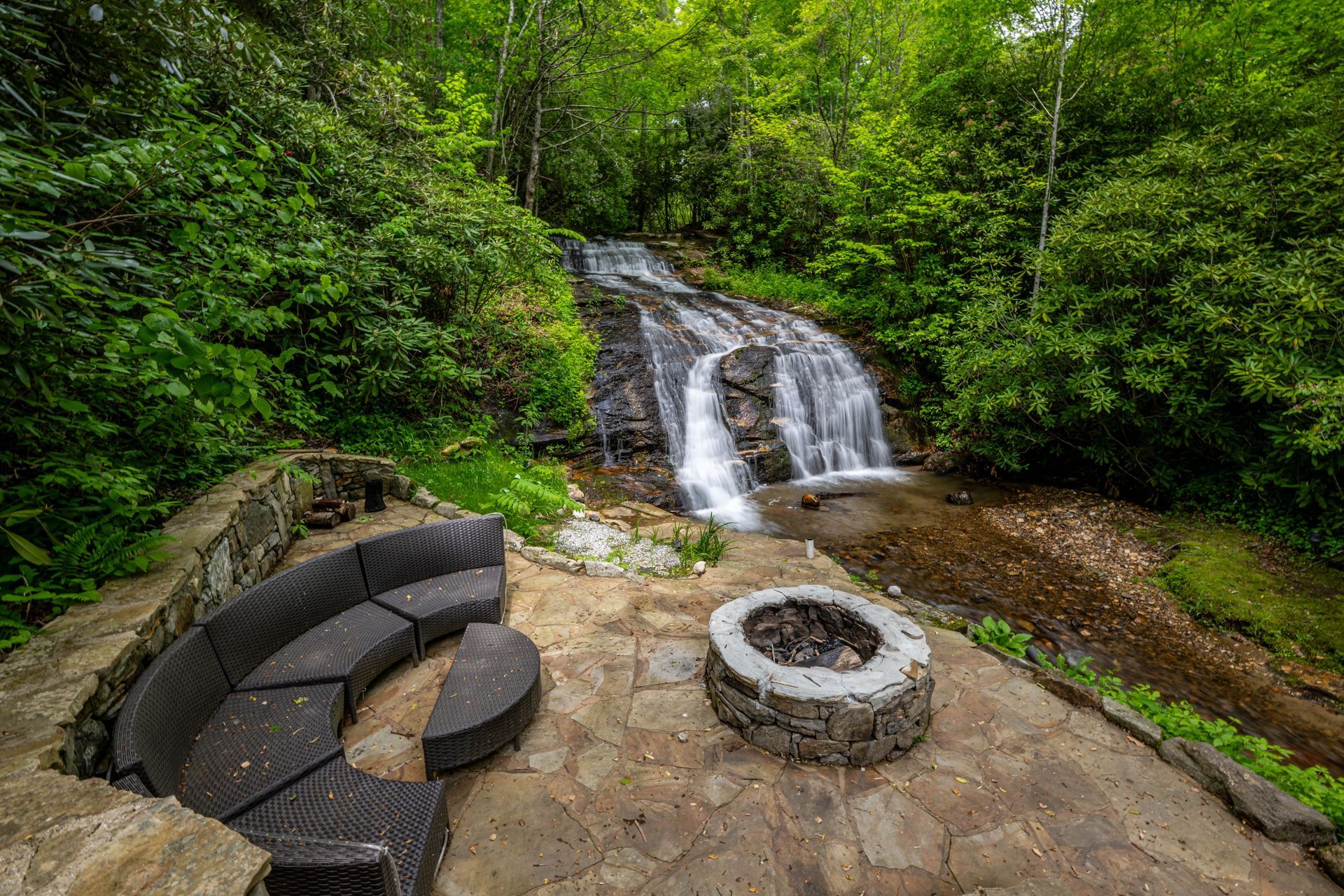 Stone patio with seating and fire pit, overlooking a waterfall in lush green forest.
