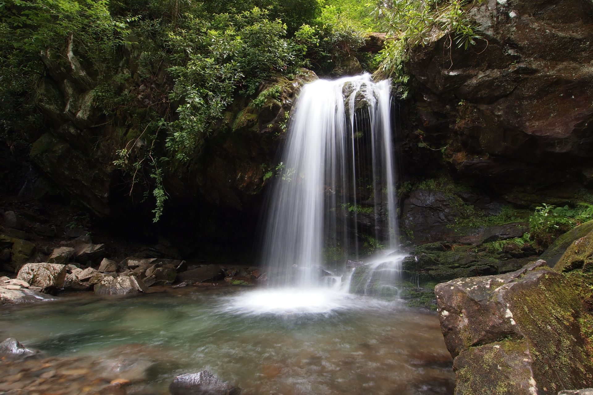 Waterfall cascading into a clear pool, surrounded by dark rocks and green foliage.