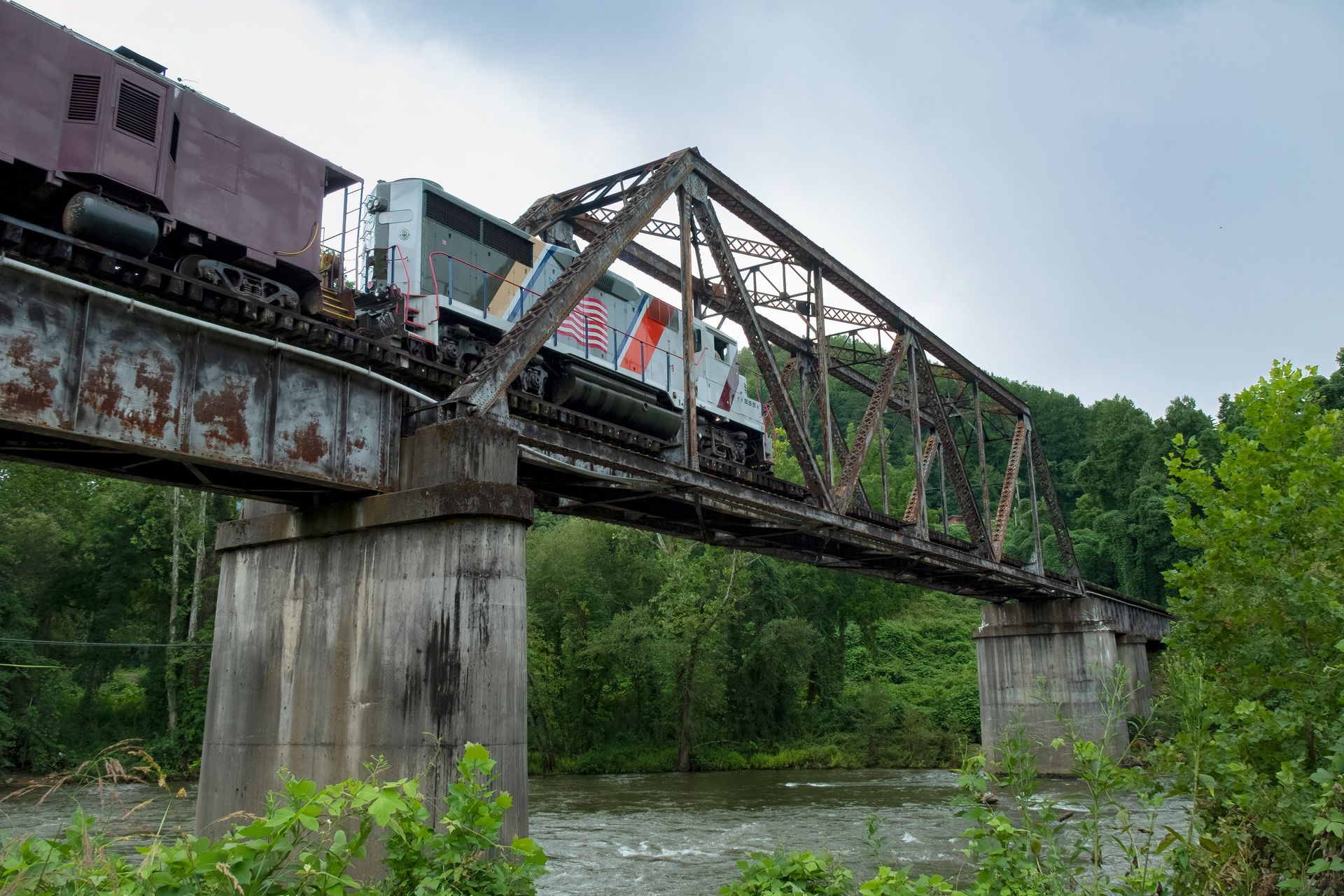 Train crossing a rusty metal bridge over a river; trees line the banks.
