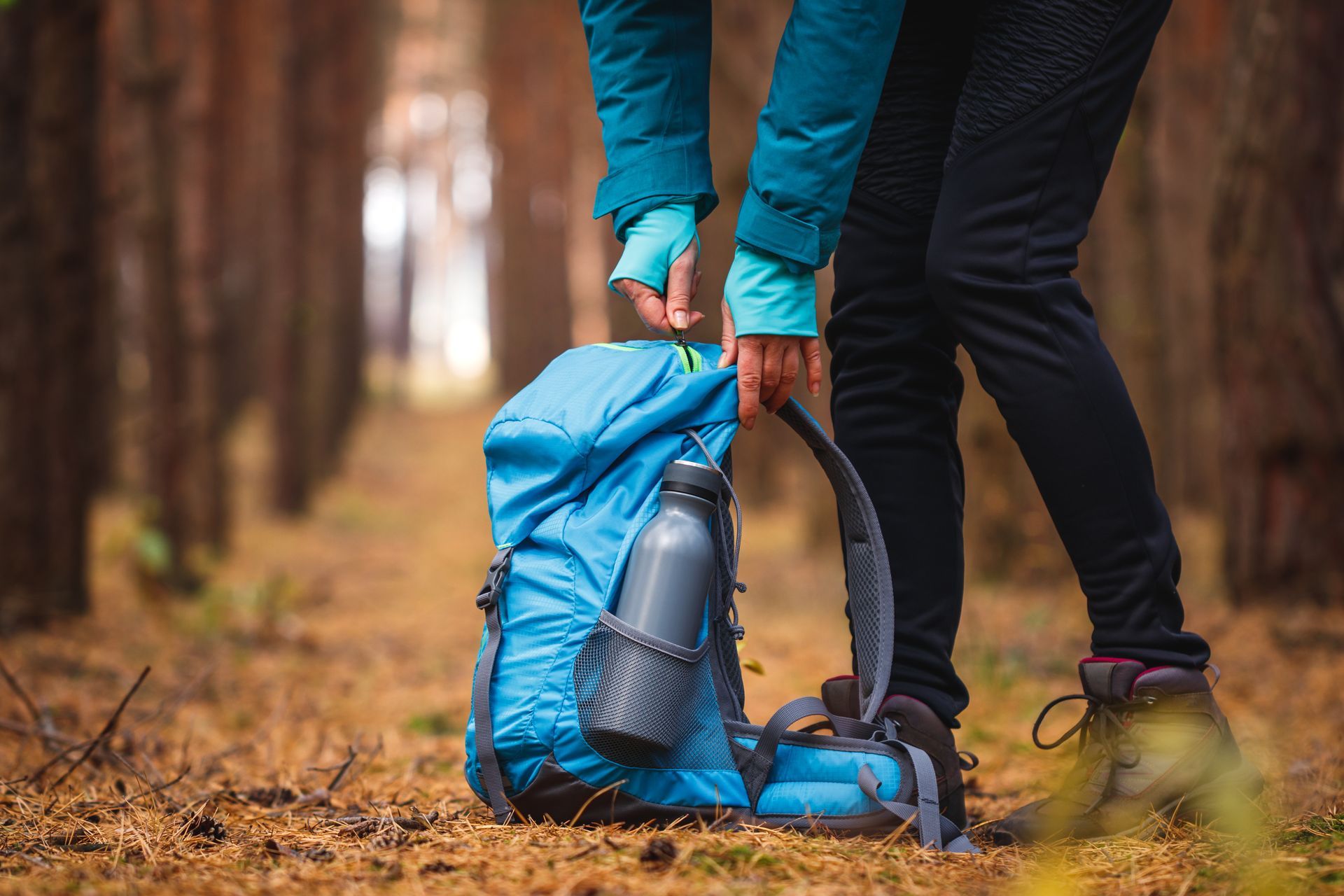 Person opening a blue backpack in a forest, water bottle in side pocket, fall leaves.