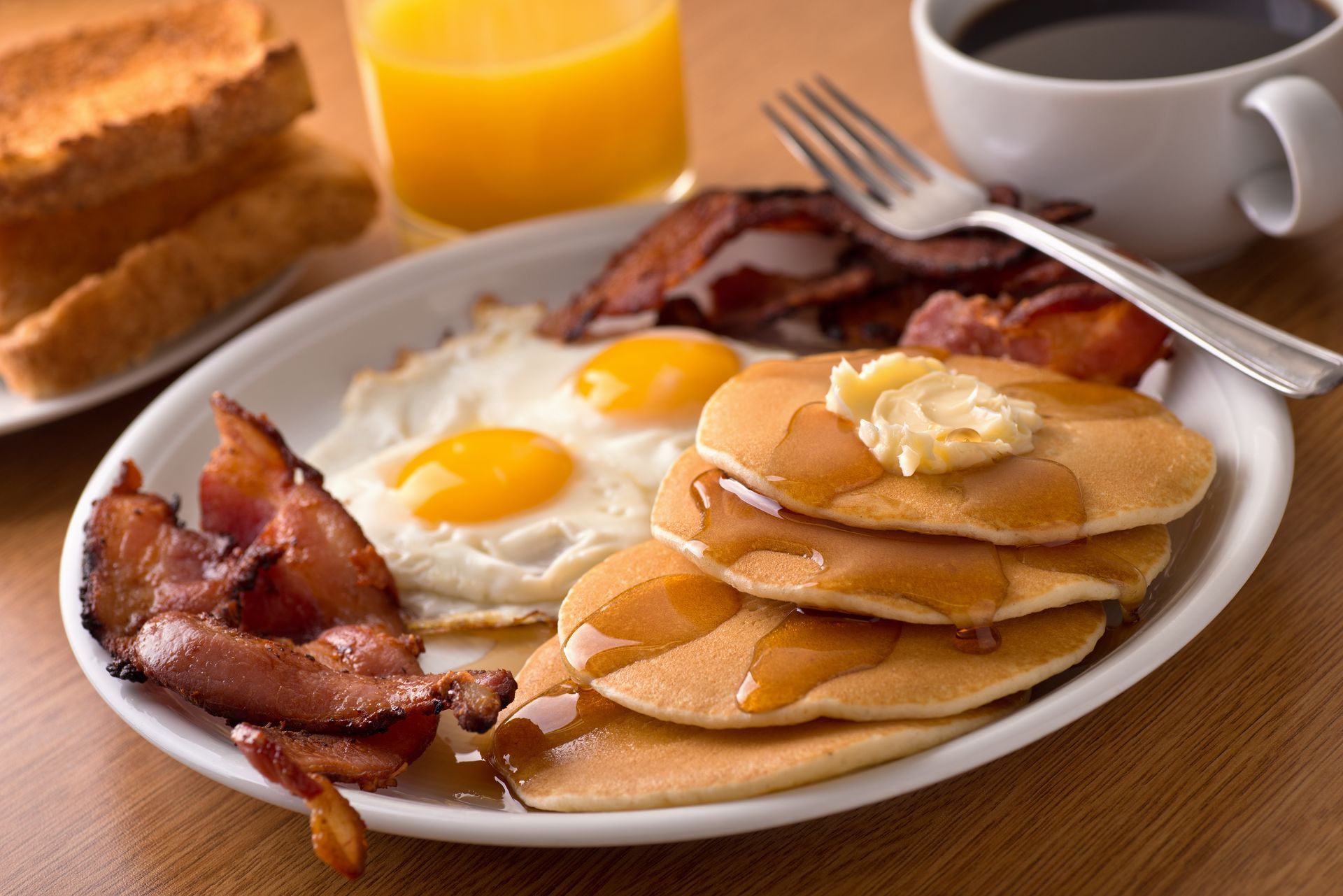 Breakfast plate with eggs, pancakes, bacon, toast, orange juice, and coffee.