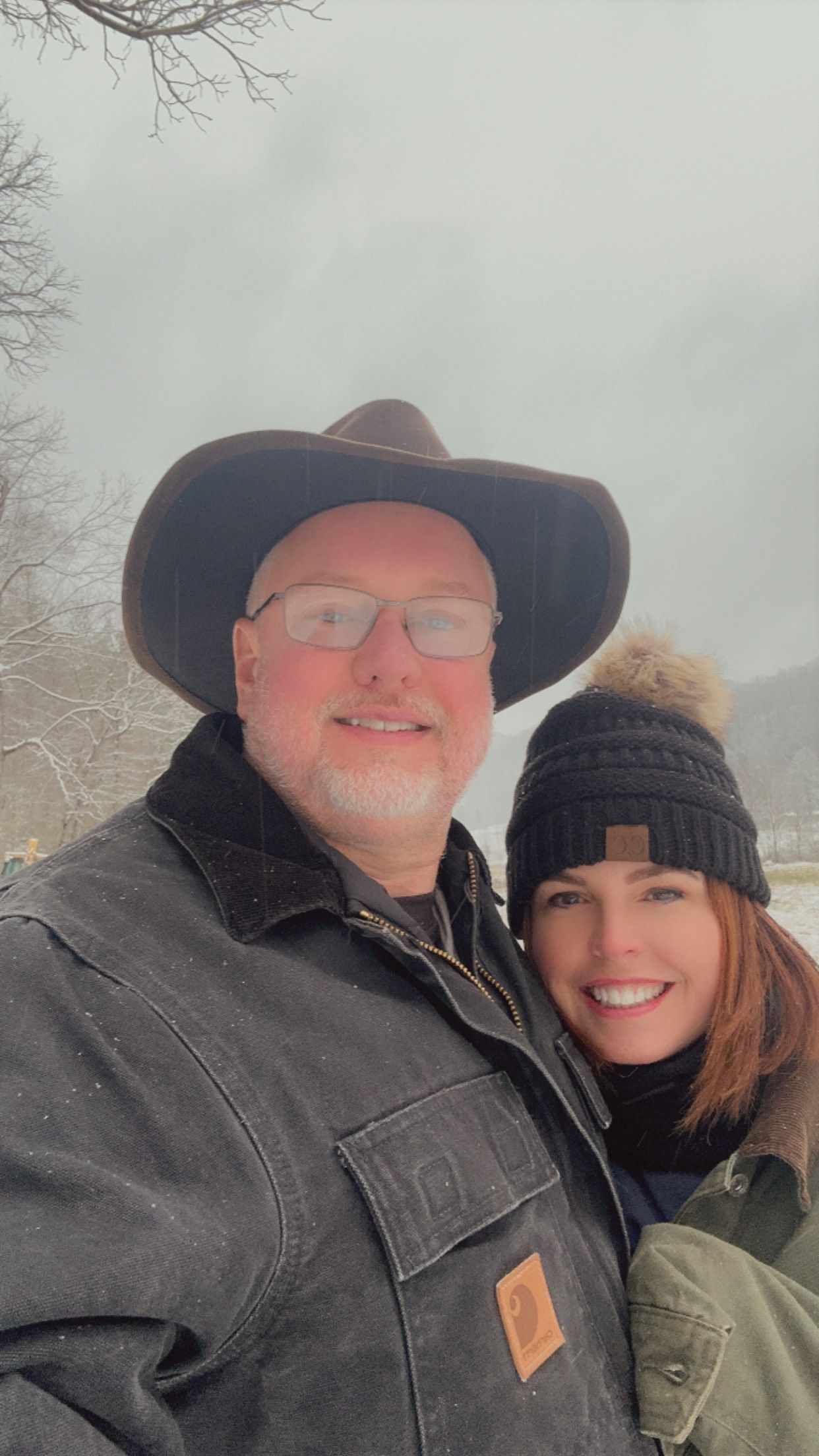 Man in cowboy hat and woman in beanie smile in a snowy landscape.