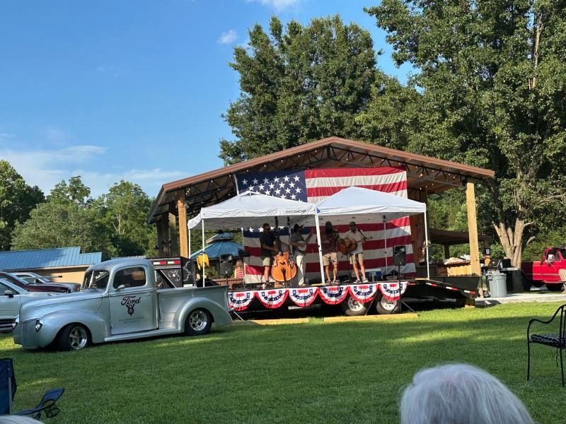 Silver truck parked near a stage with band playing under an American flag backdrop.